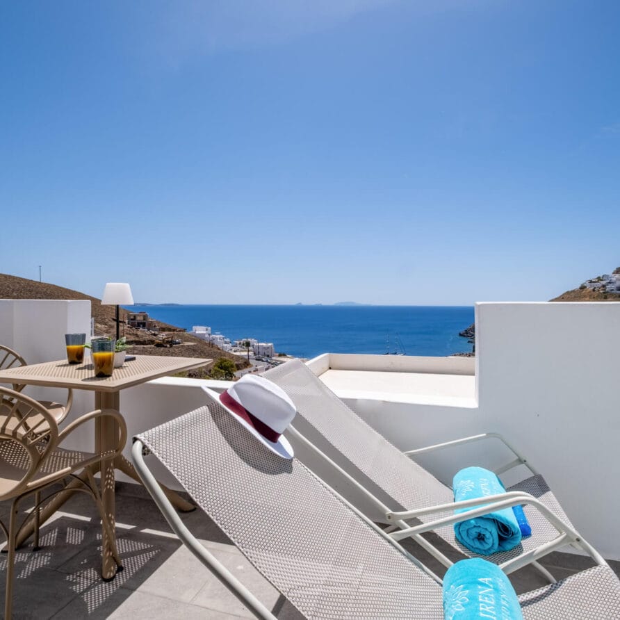 Μια βεράντα με ξαπλώστρες, ένα τραπέζι με ποτά, πετσέτες πισίνας και ένα καπέλο έχει θέα στη γαλάζια θάλασσα και τους μακρινούς λόφους κάτω από έναν καθαρό ουρανό.