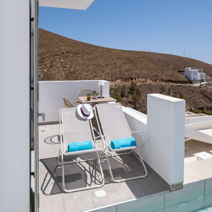 Δύο ξαπλώστρες με μπλε πετσέτες και ένα καπέλο βρίσκονται σε μια βεράντα δίπλα σε μια μικρή πισίνα, με θέα σε ένα ξηρό, λοφώδες τοπίο κάτω από έναν καταγάλανο ουρανό.