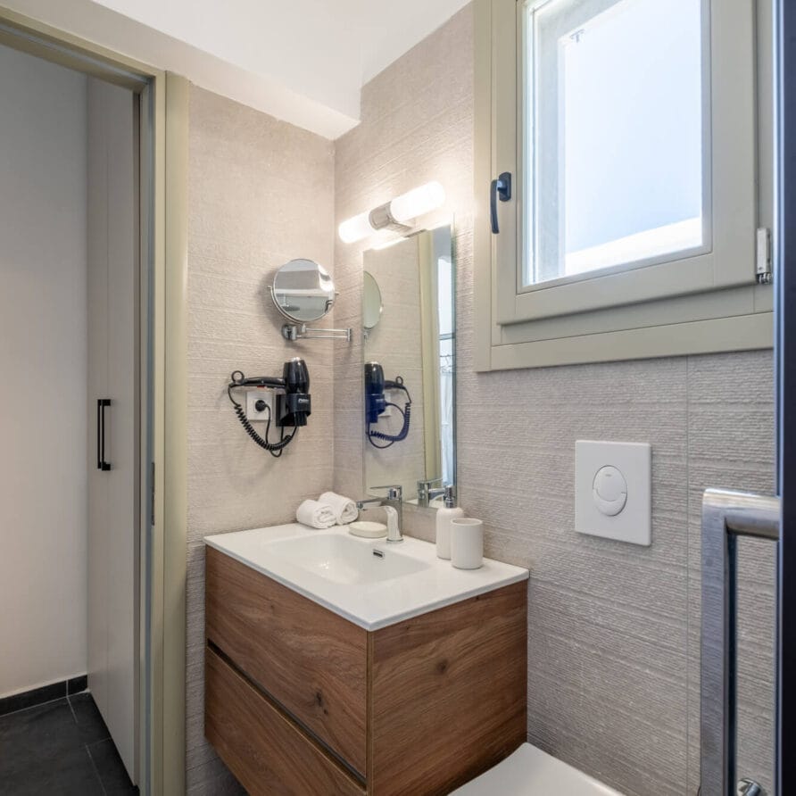 Μοντέρνο μπάνιο με ξύλινο νιπτήρα, επιτοίχιο καθρέφτη, νιπτήρα, τουαλέτα και παράθυρο. Στεγνωτήρας μαλλιών και πετσέτες τοποθετούνται κοντά στον καθρέφτη. Ουδέτερη χρωματική παλέτα.