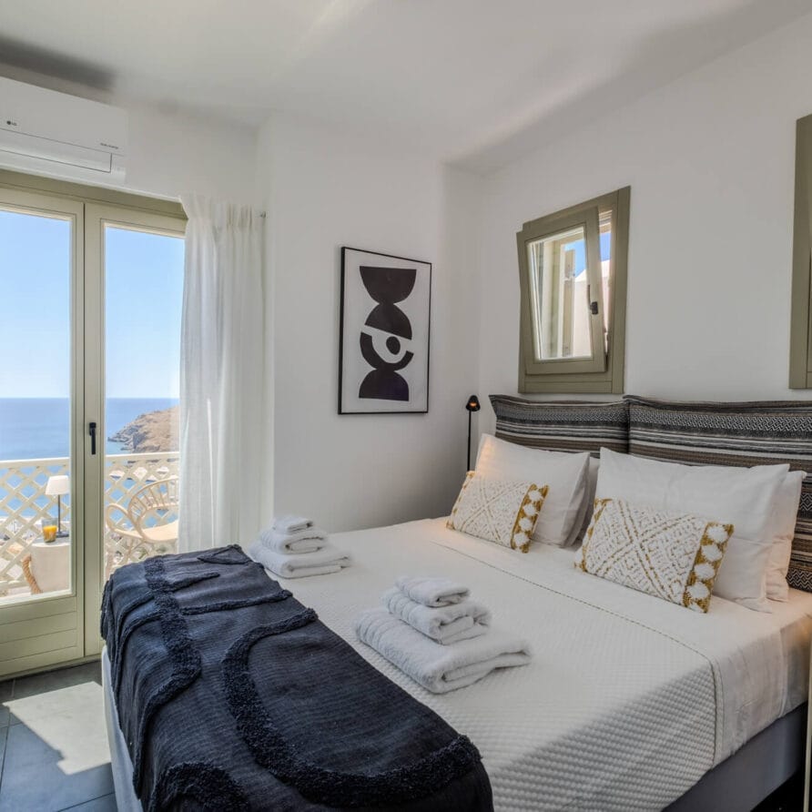 Ένα προσεγμένο διπλό κρεβάτι με μαξιλάρια και πετσέτες σε ένα φωτεινό, μοντέρνο υπνοδωμάτιο με παράθυρα και μπαλκόνι με θέα στη θάλασσα.
