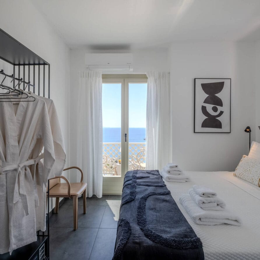 Ένα φωτεινό δωμάτιο ξενοδοχείου με ένα προσεγμένο διπλό κρεβάτι, διπλωμένες πετσέτες, μια ρόμπα σε μια ανοιχτή ντουλάπα, μια καρέκλα, έργα τέχνης στον τοίχο και μια μπαλκονόπορτα με θέα στον ωκεανό.