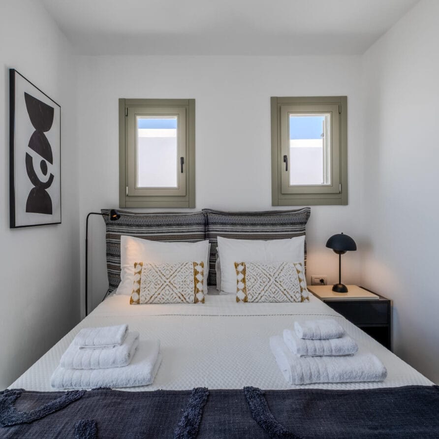 Ένα προσεγμένο κρεβάτι με δύο μαξιλάρια με σχέδια, δύο στοίβες πετσέτες, ένα μαύρο φωτιστικό σε ένα κομοδίνο, μινιμαλιστικά έργα τέχνης στον τοίχο και δύο μικρά παράθυρα πάνω από το κρεβάτι.