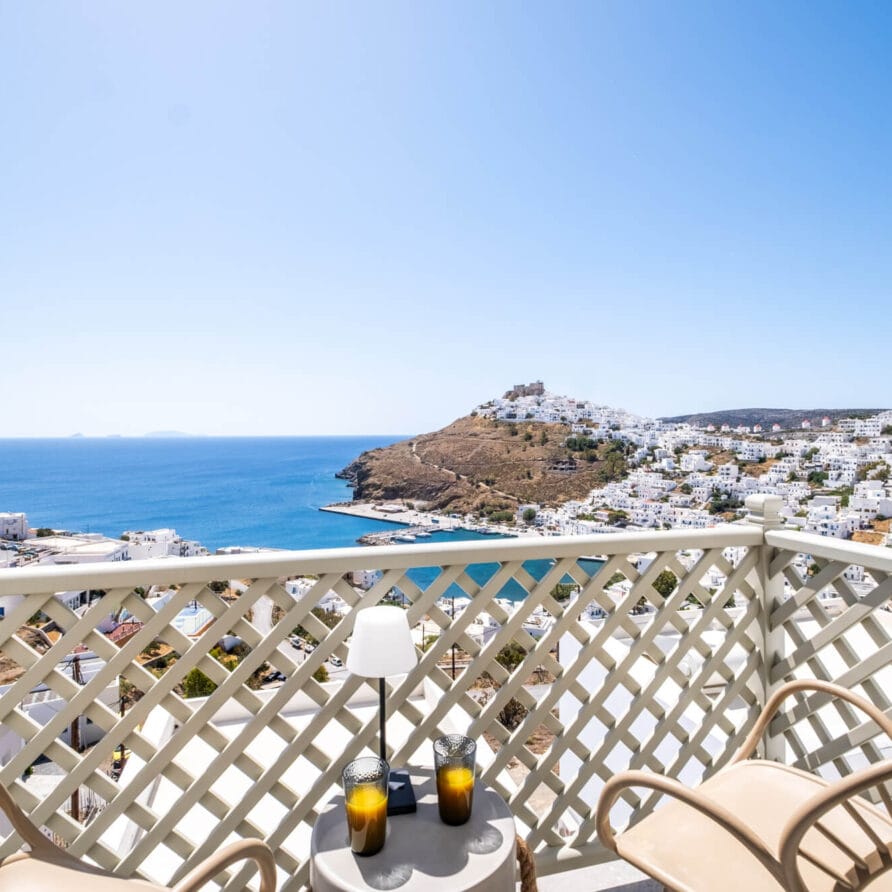 Θέα από ένα μπαλκόνι με δύο καρέκλες και ένα μικρό τραπέζι με θέα σε μια παραθαλάσσια πόλη, τη γαλάζια θάλασσα και την πλαγιά ενός λόφου κάτω από έναν καθαρό ουρανό.