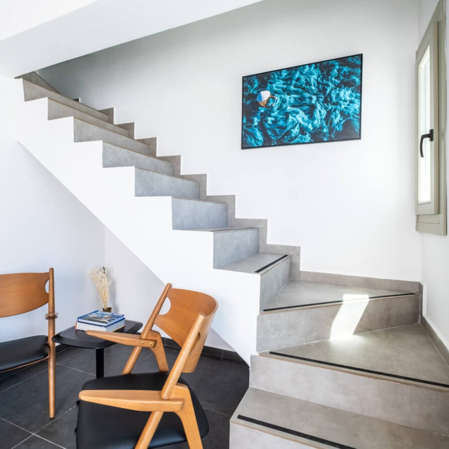 Μοντέρνα σκάλα με γκρι σκαλοπάτια, δύο ξύλινες καρέκλες και ένα μικρό τραπέζι από κάτω, αφηρημένα έργα τέχνης στον τοίχο και ένα παράθυρο που αφήνει να μπαίνει φυσικό φως.
