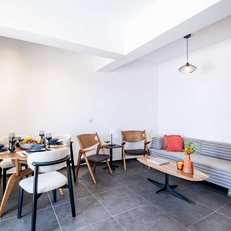 Σύγχρονος χώρος καθιστικού και τραπεζαρίας με σετ τραπεζαρίας, τέσσερις καρέκλες, έναν καναπέ με σχέδια, δύο πολυθρόνες, ένα τραπεζάκι σαλονιού και μινιμαλιστική διακόσμηση σε γκρι πλακάκια δαπέδου.