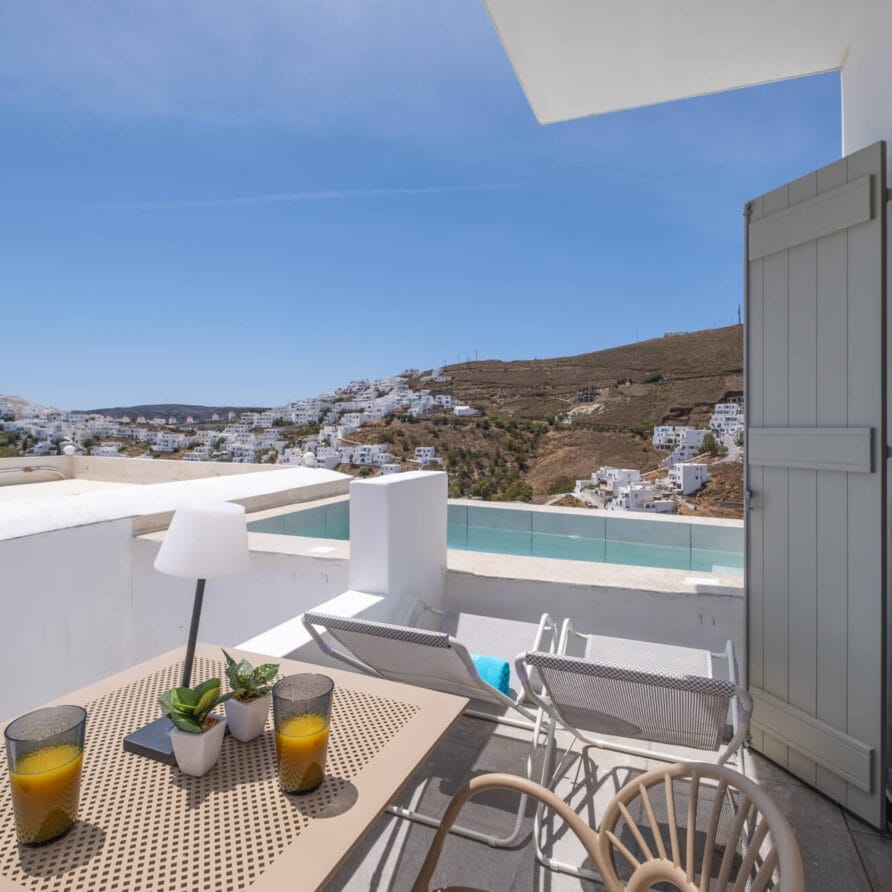 Ένα μπαλκόνι με τραπέζι, καρέκλες και δύο πορτοκαλί ποτά έχει θέα σε μια πόλη στην πλαγιά ενός λόφου με λευκά κτίρια κάτω από έναν καταγάλανο ουρανό.