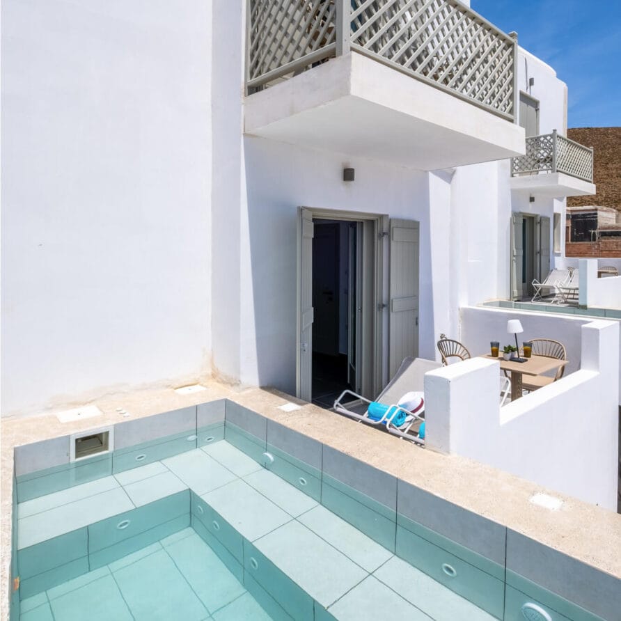 Ένα μοντέρνο λευκό κτίριο με μπαλκόνια και μια μικρή, ορθογώνια πισίνα με γυάλινους τοίχους στο προσκήνιο κάτω από έναν καταγάλανο ουρανό.