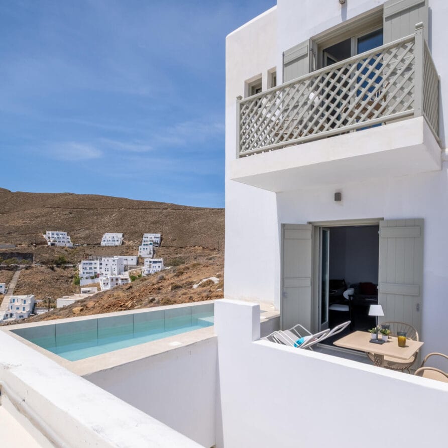 Ένα λευκό μοντέρνο κτίριο με ανοιχτές μπαλκονόπορτες έχει θέα σε μια πισίνα και μια πλαγιά λόφου γεμάτη με παρόμοιες λευκές κατασκευές κάτω από έναν καταγάλανο ουρανό.