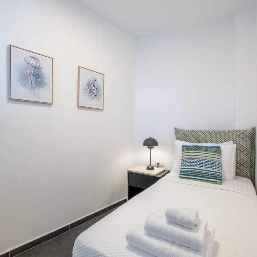 Μινιμαλιστικό υπνοδωμάτιο με μονό κρεβάτι, προσεγμένα διπλωμένες πετσέτες, κομοδίνο με φωτιστικό και δύο πλαισιωμένες εκτυπώσεις μεδουσών σε λευκό τοίχο.