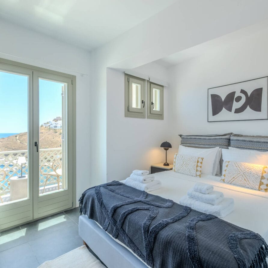 Φωτεινό υπνοδωμάτιο με διπλό κρεβάτι, τακτοποιημένες πετσέτες, δύο παράθυρα, μπαλκονόπορτα με θέα στη θάλασσα και μινιμαλιστική διακόσμηση που περιλαμβάνει μια αφηρημένη τέχνη τοίχου πάνω από το κεφαλάρι του κρεβατιού.