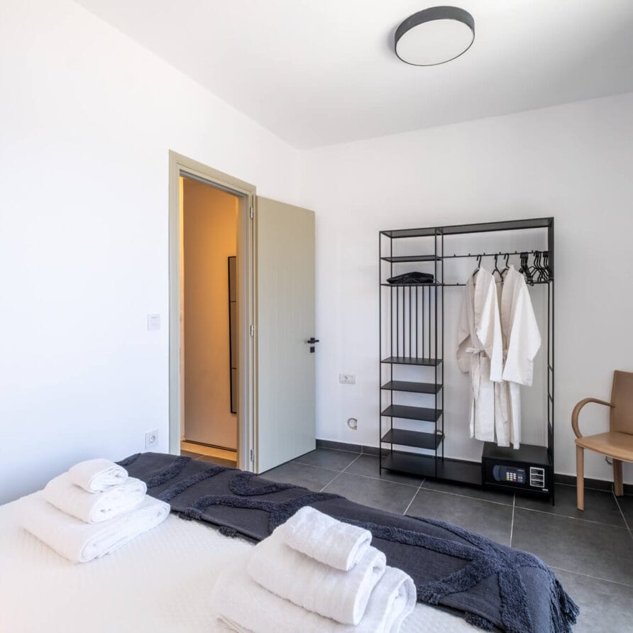 Μινιμαλιστικό υπνοδωμάτιο με κρεβάτι, διπλωμένες πετσέτες, ανοιχτή ντουλάπα με μπουρνούζια και κρεμάστρες, καρέκλα και φυσικό φως από παράθυρο.