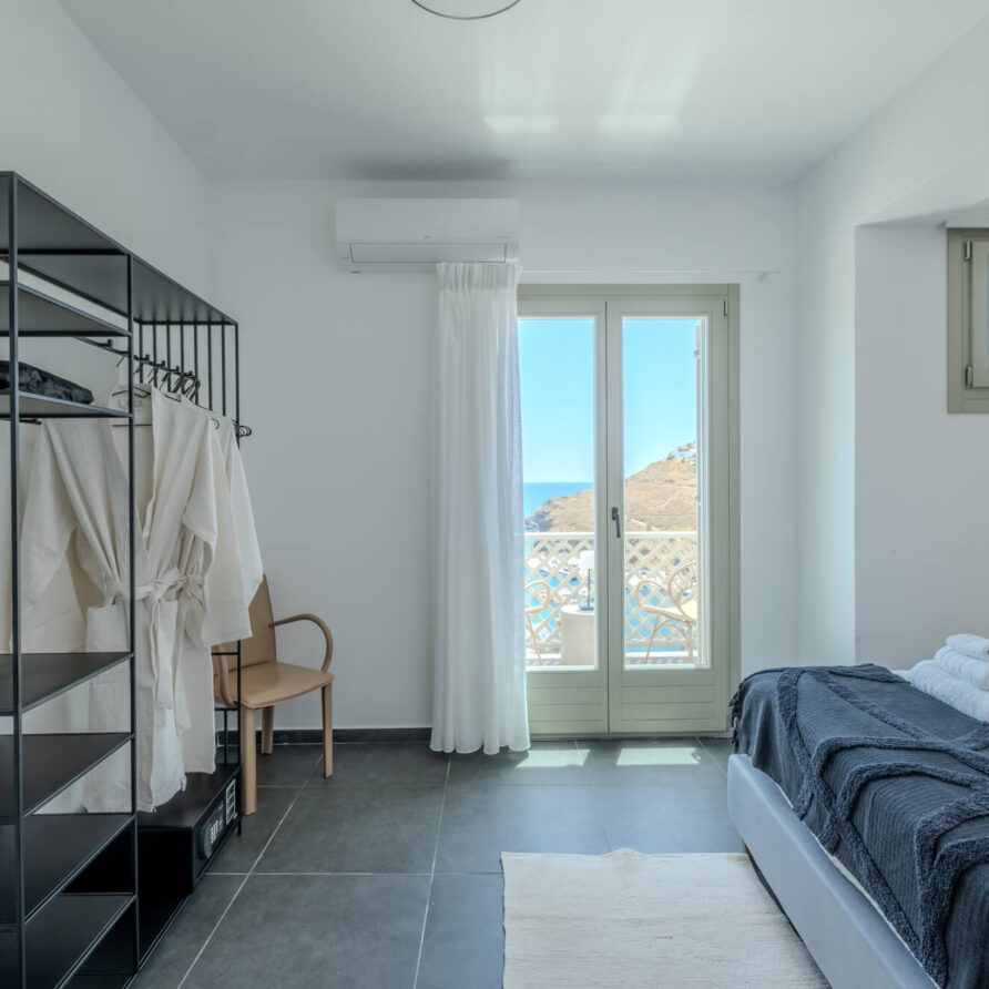 Μινιμαλιστικό υπνοδωμάτιο με ανοιχτή ντουλάπα, μπουρνούζια, καρέκλα, στρωμένο κρεβάτι με σκούρα μπλε κουβέρτα, διπλωμένες πετσέτες και παράθυρο που βλέπει σε μπαλκόνι και θέα στη θάλασσα.
