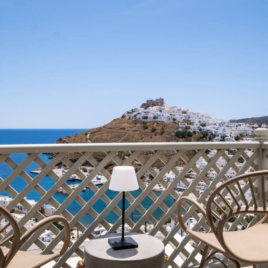 Δύο καρέκλες και ένα μικρό τραπέζι με μια λάμπα σε ένα μπαλκόνι έχουν θέα σε μια πόλη στην κορυφή ενός λόφου και στη θάλασσα κάτω από έναν καταγάλανο ουρανό.