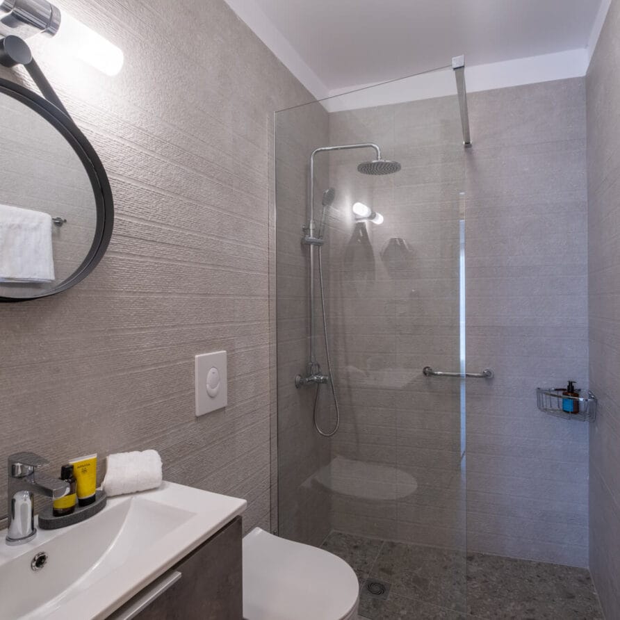 Μοντέρνο μπάνιο με επιτοίχιο νιπτήρα, στρογγυλό καθρέφτη, στεγνωτήρα μαλλιών, γυάλινη καμπίνα ντους, κρεμάστρα για πετσέτες και γκρι ανάγλυφους τοίχους.