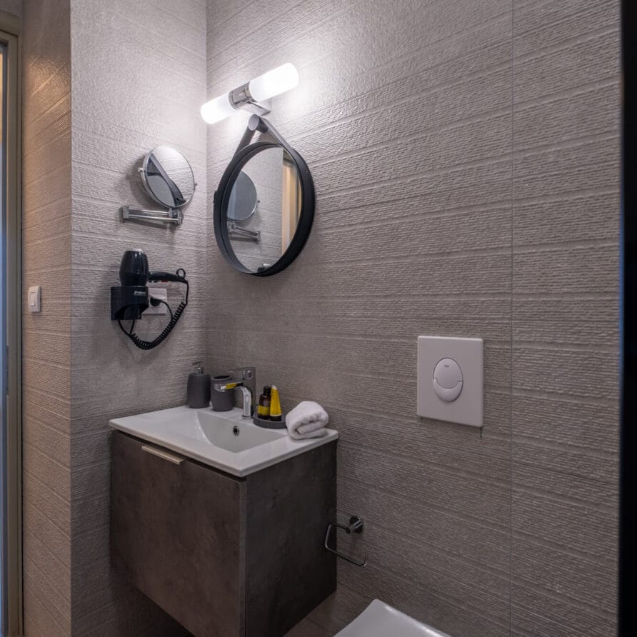 Μοντέρνο μπάνιο με γκρι πλακάκια στους τοίχους, επιτοίχιο νιπτήρα και νιπτήρα, στρογγυλό καθρέφτη με φωτιστικό από πάνω και τουαλέτα σε πρώτο πλάνο.