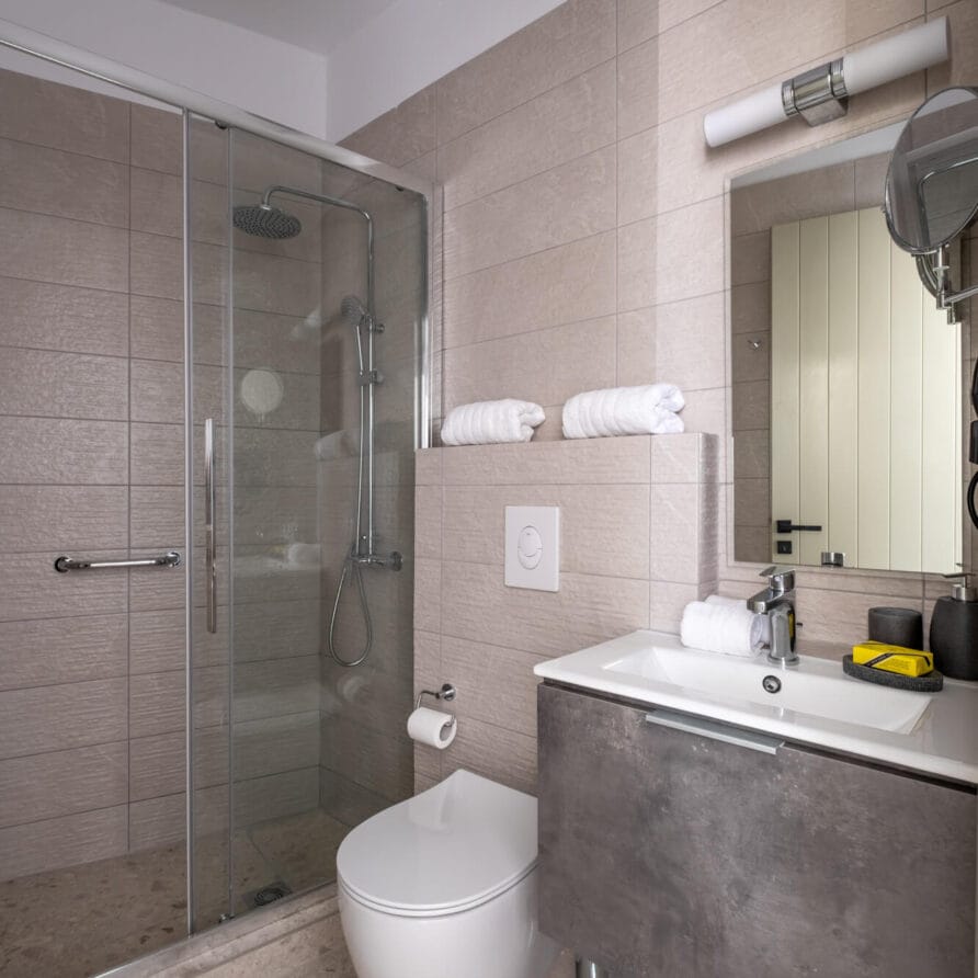 Μοντέρνο μπάνιο με γυάλινη καμπίνα ντους, επιτοίχια τουαλέτα, νιπτήρα με ντουλάπι, καθρέφτη, κρεμάστρες για πετσέτες και τακτοποιημένα διπλωμένες λευκές πετσέτες.