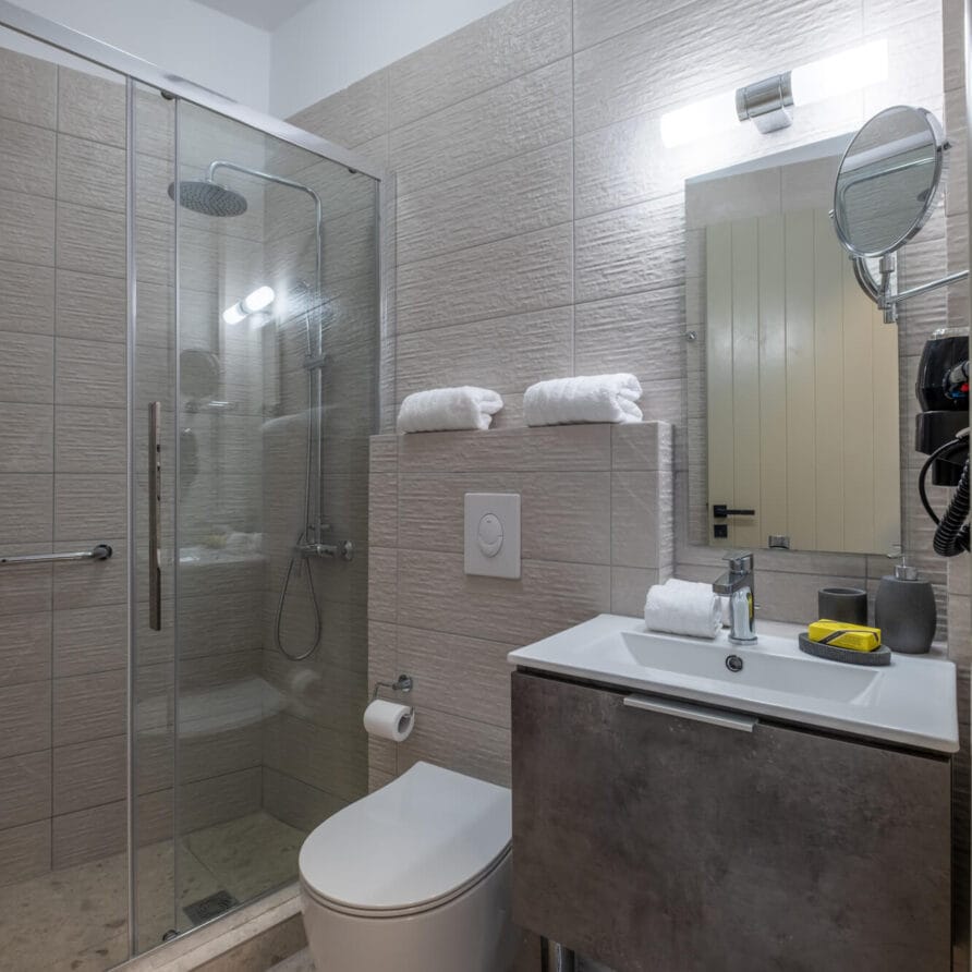 Μοντέρνο μπάνιο με γυάλινη καμπίνα ντους, επιτοίχια τουαλέτα, νιπτήρα με ντουλάπι, καθρέφτη και δύο διπλωμένες πετσέτες σε ένα περβάζι πάνω από την τουαλέτα.