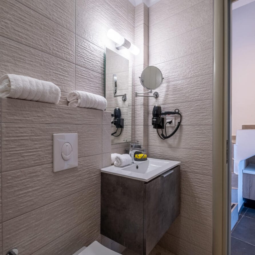 Μοντέρνο μπάνιο με μπεζ πλακάκια στους τοίχους, επιτοίχια τουαλέτα, νιπτήρα που αιωρείται, καθρέφτη, επιτοίχιο στεγνωτήρα μαλλιών, πετσέτες και θήκη για χαρτί υγείας.