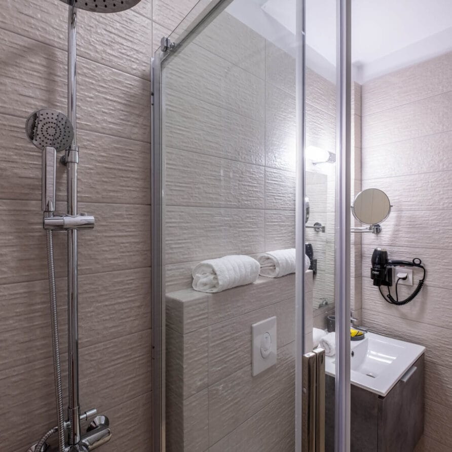 Μικρό μοντέρνο μπάνιο με γυάλινη καμπίνα ντους, επιτοίχια κεφαλή ντους, νιπτήρα με καθρέφτη, στεγνωτήρα μαλλιών και πετσέτες τοποθετημένες τακτοποιημένα σε ράφι.