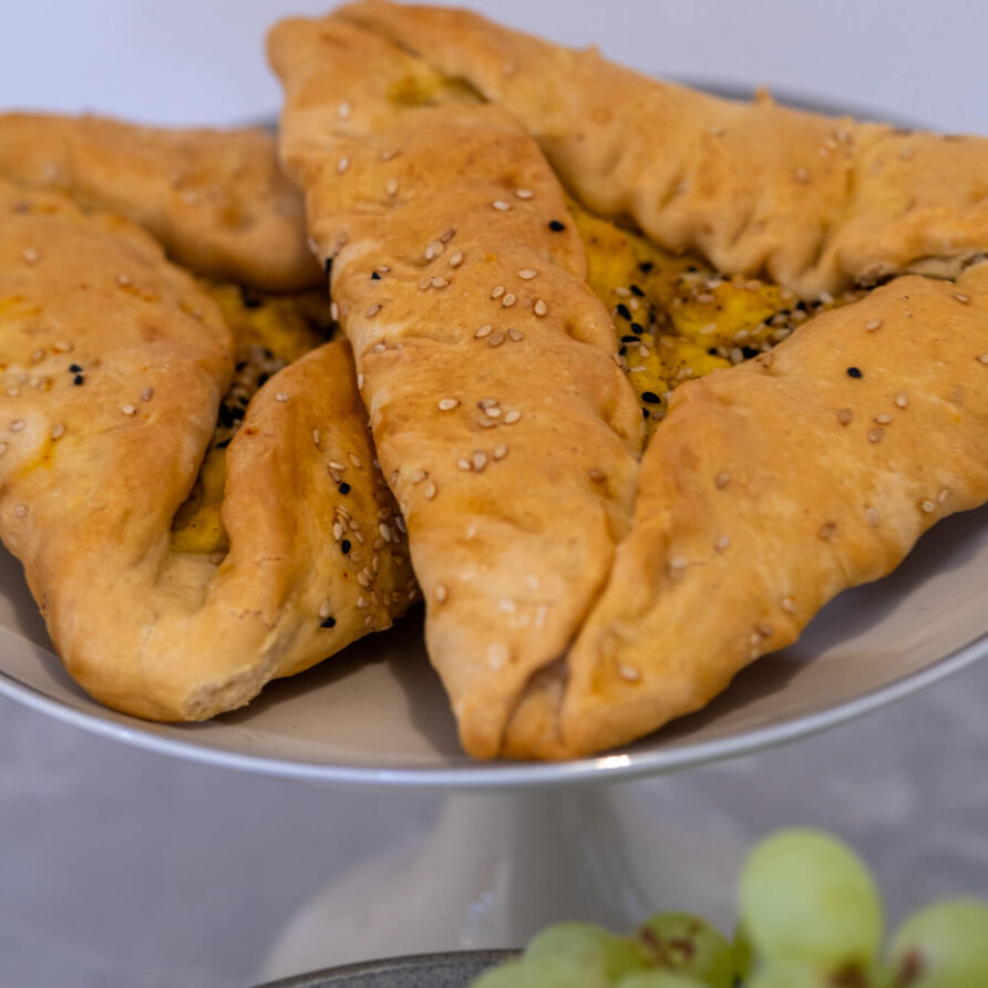 Τρία κομμάτια ζύμης με σουσάμι και αλμυρή γέμιση είναι τοποθετημένα σε ένα πιάτο, με ένα μικρό τσαμπί πράσινα σταφύλια ορατό στο προσκήνιο.