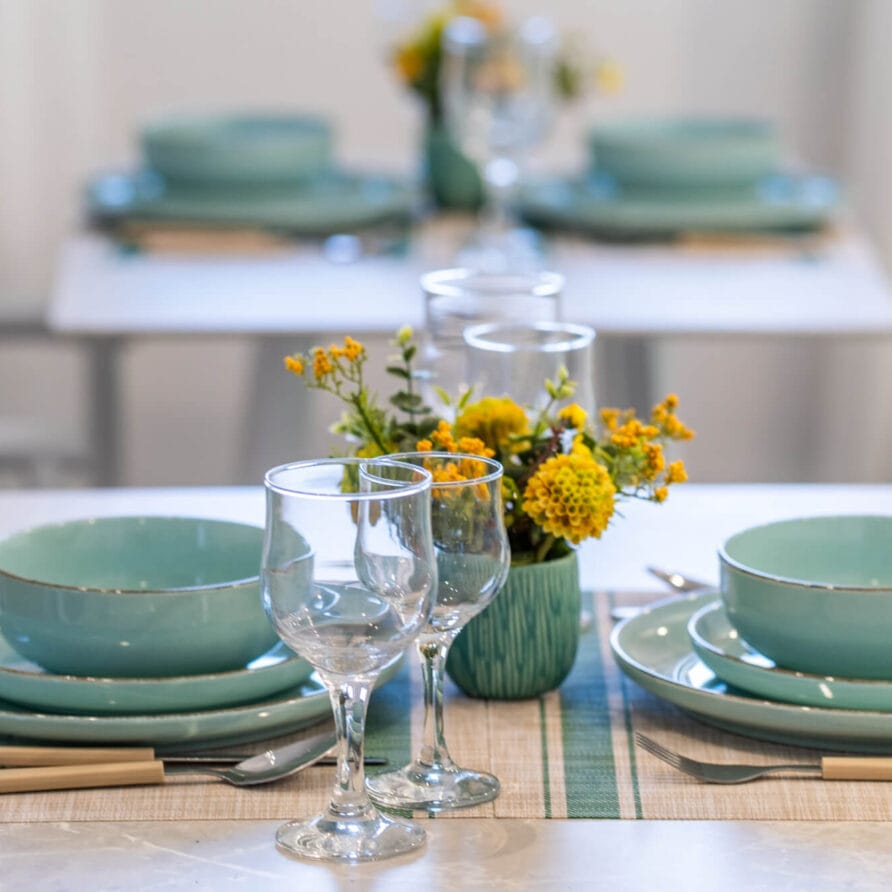 Ένα όμορφα στρωμένο τραπέζι φαγητού με τιρκουάζ πιάτα, ποτήρια κρασιού και κεντρικά διακοσμητικά με κίτρινα λουλούδια σε σουπλά, με ένα άλλο παρόμοια στρωμένο τραπέζι στο βάθος.