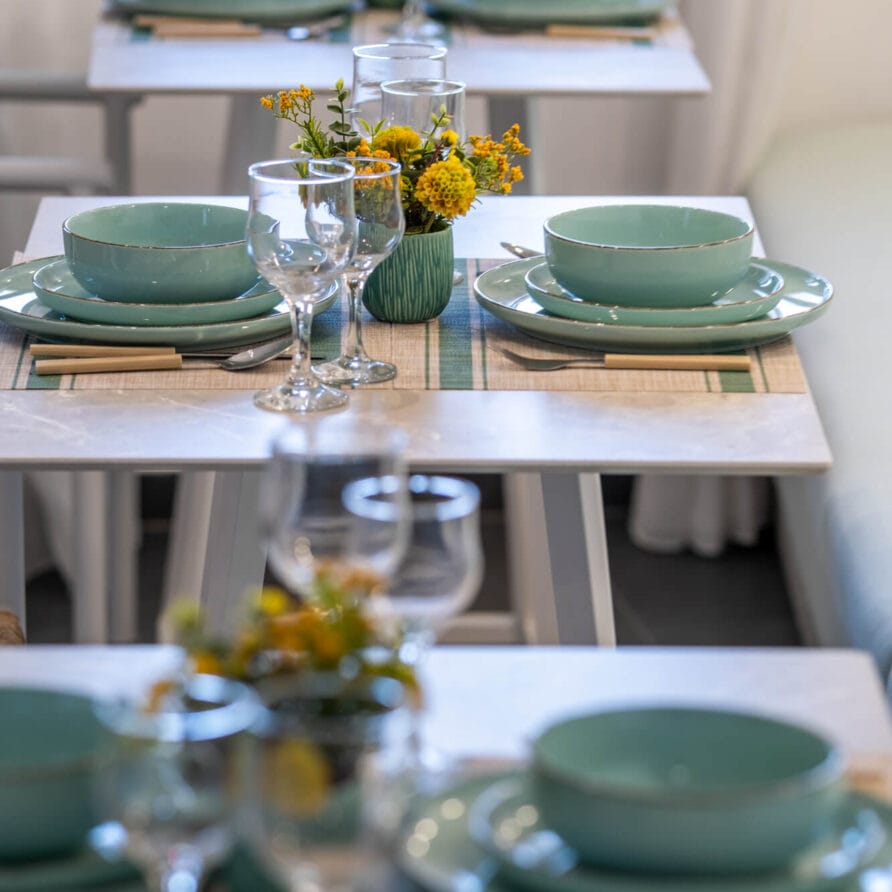 Τραπέζια στρωμένα με τιρκουάζ πιάτα, ποτήρια κρασιού και κίτρινα φλοράλ διακοσμητικά στοιχεία, τοποθετημένα σε μια τακτοποιημένη σειρά.