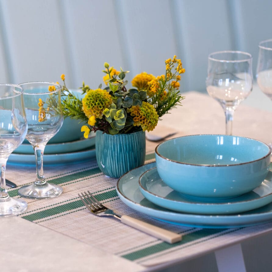 Ένα τραπέζι στρωμένο για δύο με τιρκουάζ πιάτα, ασημένια μαχαιροπίρουνα, τέσσερα ποτήρια κρασιού και ένα μικρό βάζο με κίτρινα λουλούδια σε ένα ριγέ σουπλά.