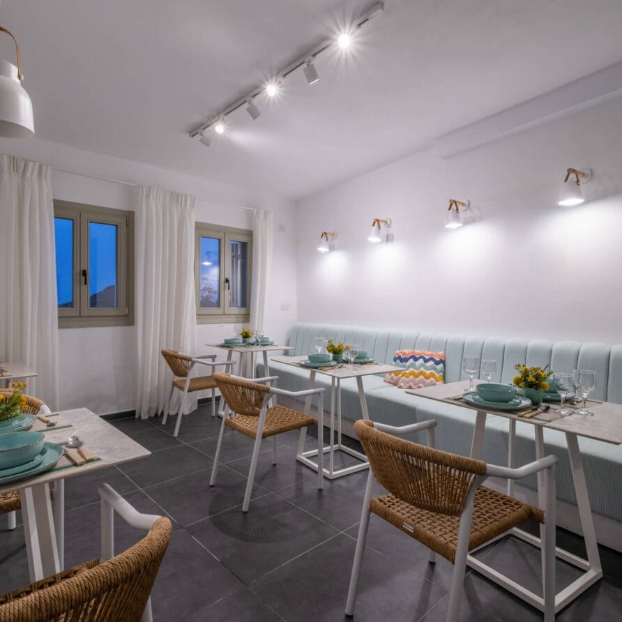 Μικρή, μοντέρνα τραπεζαρία με ανοιχτόχρωμους μπλε πάγκους, ψάθινες καρέκλες, λευκά τραπέζια στρωμένα με μπλε πιάτα και διακοσμητικά κίτρινα λουλούδια σε κάθε τραπέζι.