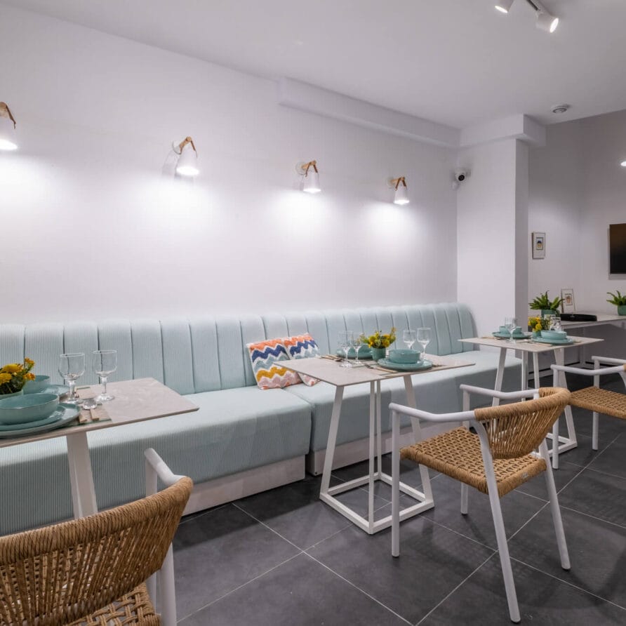 Μοντέρνα τραπεζαρία με ανοιχτό μπλε παγκάκι με επένδυση, ψάθινες καρέκλες, μικρά τραπέζια στρωμένα με πιάτα, γλάστρες με φυτά και επιτοίχια τηλεόραση με έντονο λευκό φωτισμό.