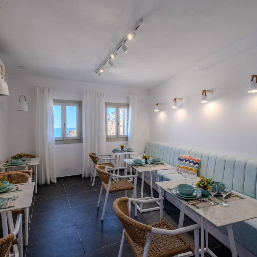 Μια μικρή, μοντέρνα τραπεζαρία με ανοιχτόχρωμα μπλε καθίσματα, ψάθινες καρέκλες, λευκά τραπέζια στρωμένα για γεύματα και παράθυρα που αφήνουν να μπαίνει φυσικό φως.