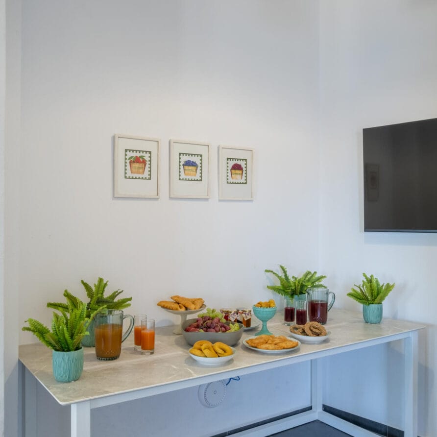 Ένα λευκό τραπέζι με ποτά, φρούτα, σνακ και φυτά σε γλάστρες είναι τοποθετημένο δίπλα σε έναν λευκό τοίχο, κάτω από κορνιζαρισμένες φωτογραφίες και μια τηλεόραση.