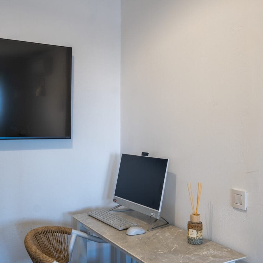 Ένας μικρός χώρος εργασίας με οθόνη, πληκτρολόγιο και ποντίκι σε ένα γκρι γραφείο, δίπλα σε έναν διαχύτη και μια ψάθινη καρέκλα. Μια τηλεόραση τοποθετημένη στον τοίχο είναι ορατή στα αριστερά.