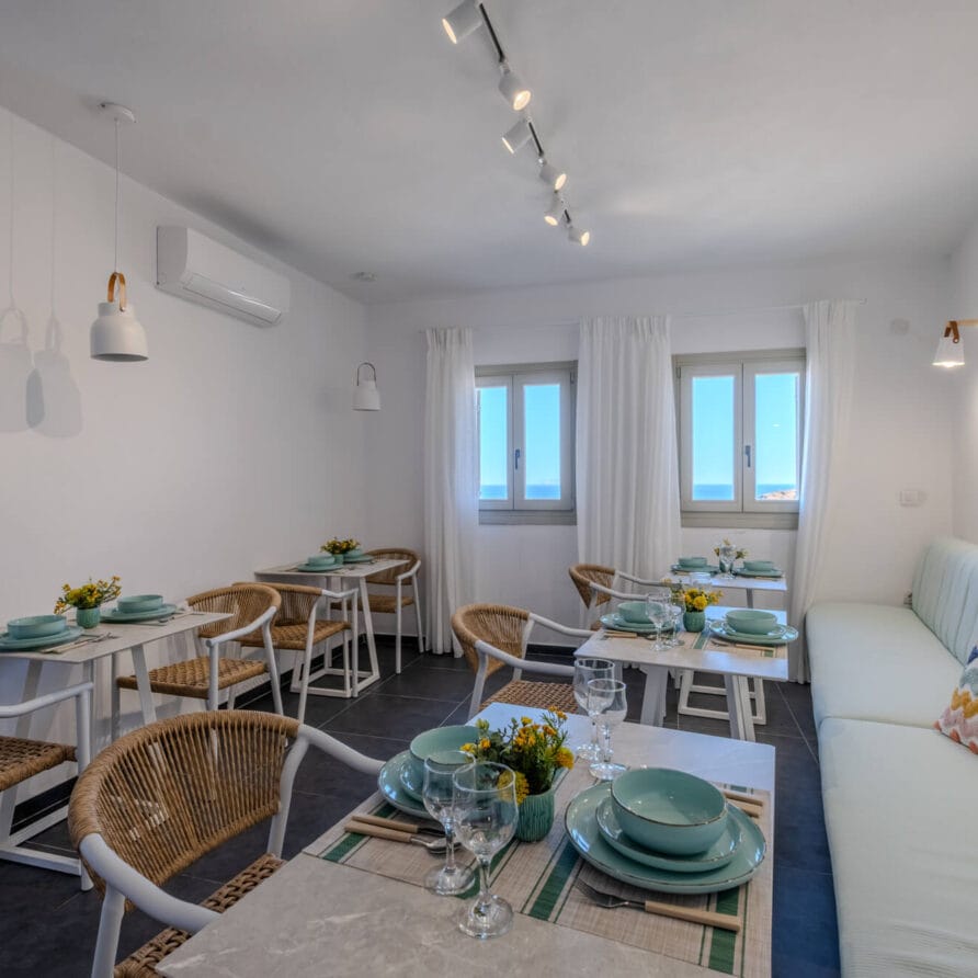 Φωτεινή τραπεζαρία με πολλά τραπέζια, ψάθινες καρέκλες, ένα μακρύ παγκάκι με μαλακά μαξιλάρια, σετ σερβίτσια και παράθυρα που αφήνουν να μπαίνει φυσικό φως.